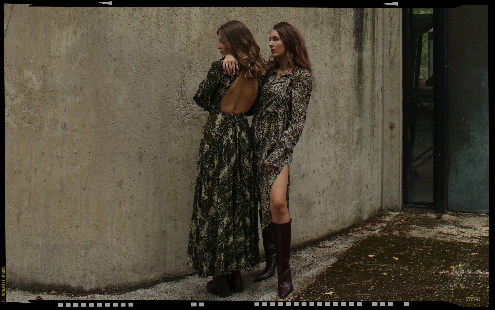 Zwei Frauen in Maxikleidern mit Schlangenprint und gedecktem Grünton lehnen an einer Betonwand – herbstlicher Secondhand-Look mit 70s-Flair, fließenden Stoffen und kniehohen Stiefeln. Fotografiert im natürlichen Licht für eine urbane, moderne Ästhetik