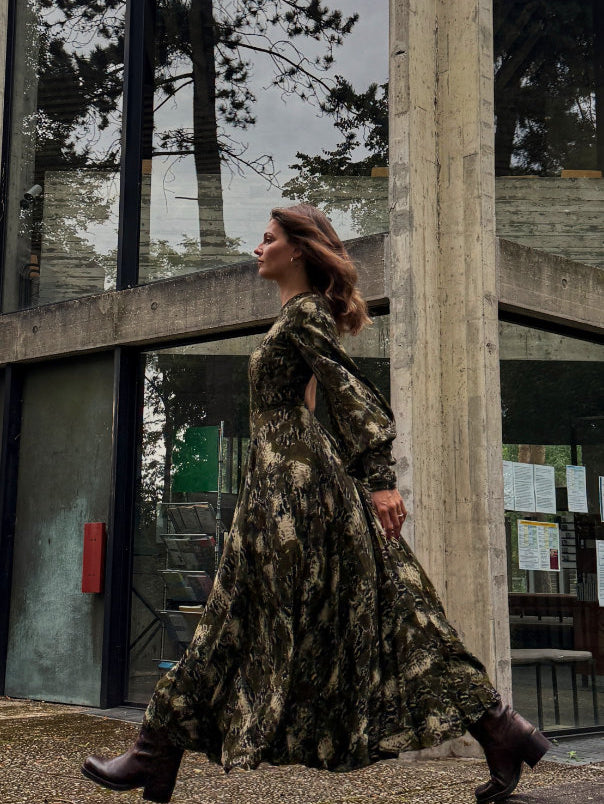 Frau trägt langes olivgrünes Maxikleid mit Print von Rotate Birger Christensen und Stiefel vor moderner Betonarchitektur – nachhaltiger Secondhand-Look