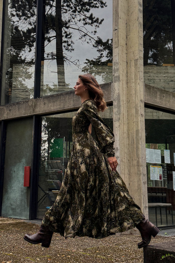 Frau trägt langes olivgrünes Maxikleid mit Print von Rotate Birger Christensen und Stiefel vor moderner Betonarchitektur – nachhaltiger Secondhand-Look